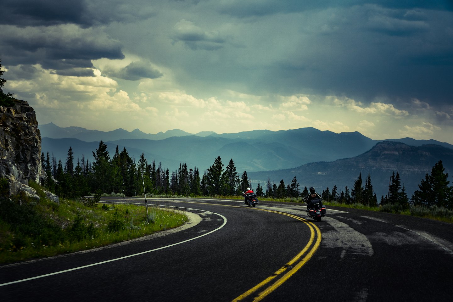 Due motociclisti su una strada tortuosa tra pini verdi.